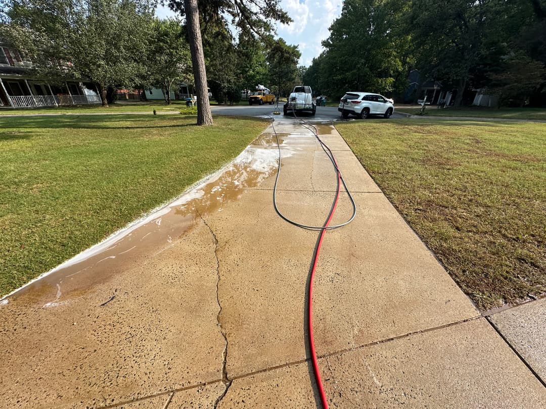 Sidewalk being cleaned with water and hose, with vehicles and trees in the background.