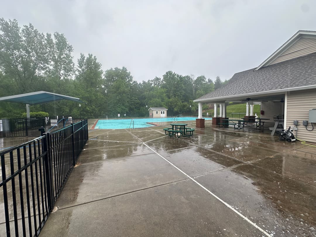 Empty outdoor swimming pool area on a rainy day with picnic tables and a nearby building.