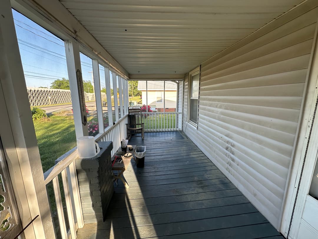 Spacious porch with wooden flooring and windows, featuring a small table and plants outside.