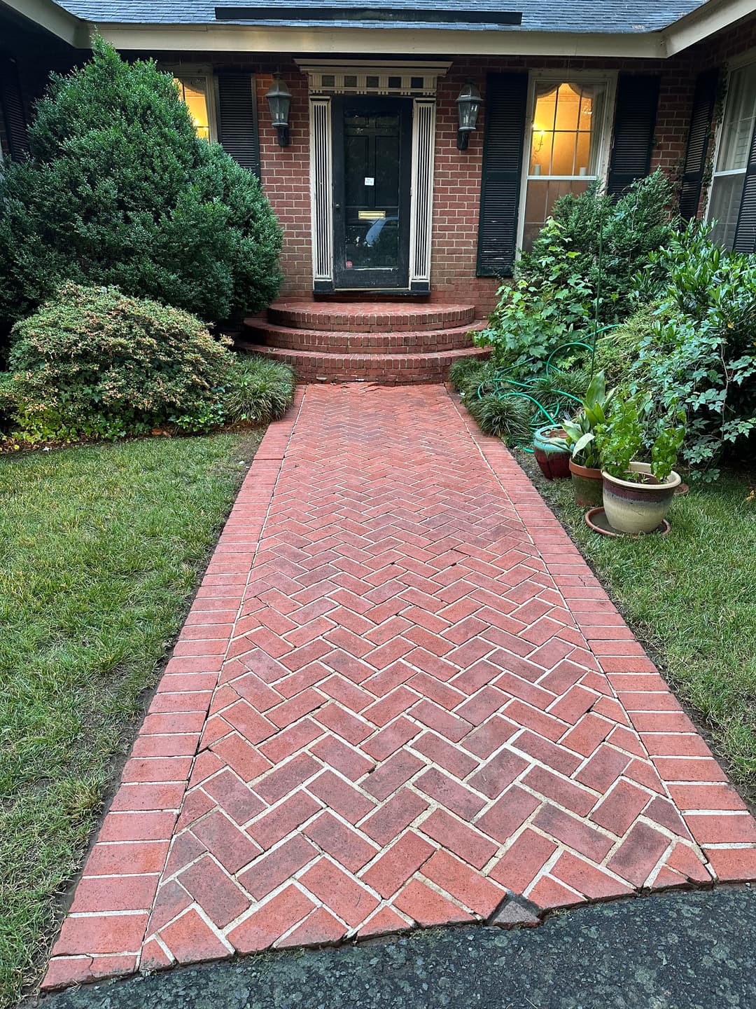 Brick walkway leading to a home's front porch, surrounded by greenery and potted plants.