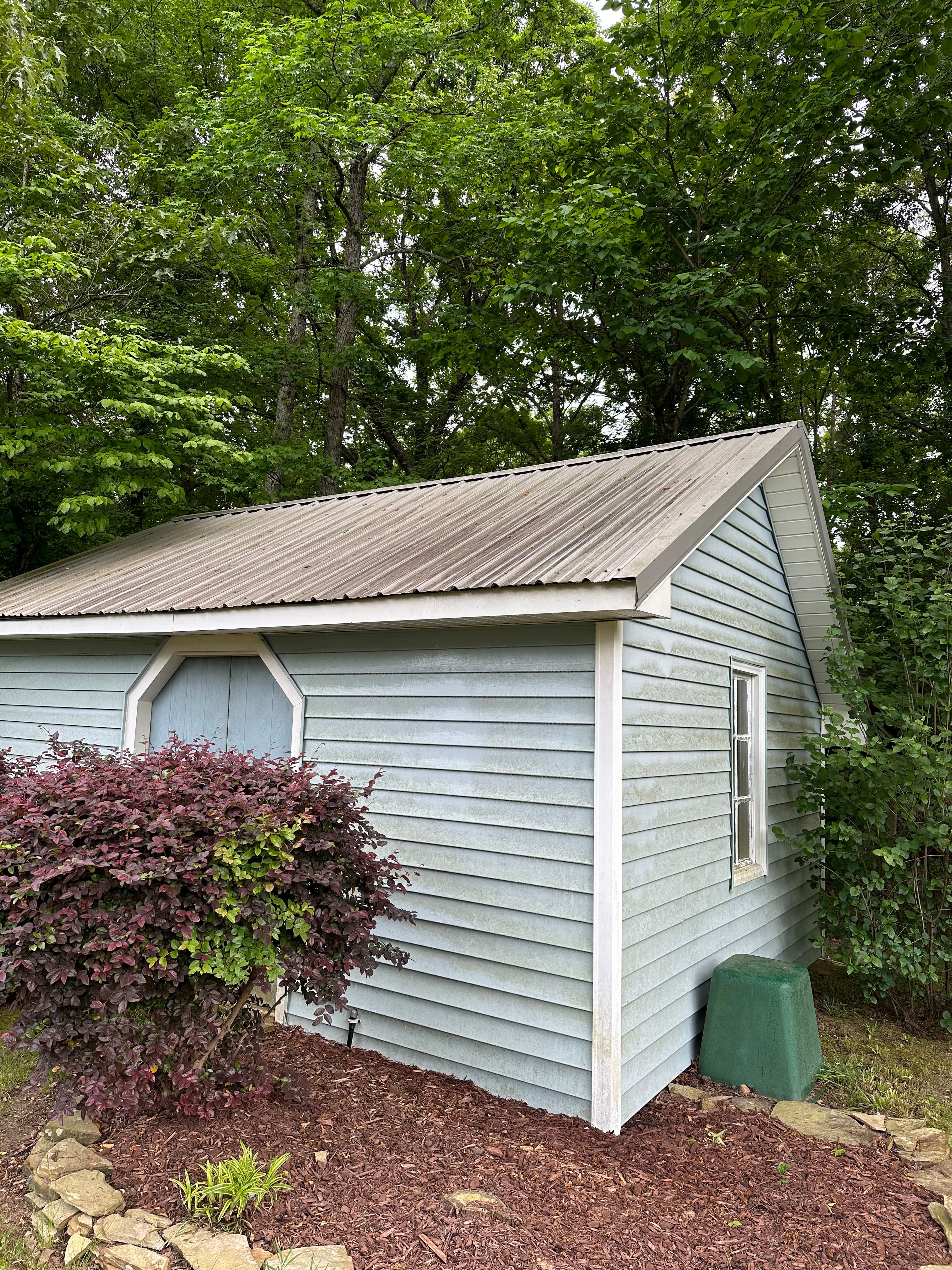 Small light blue storage shed surrounded by green trees and landscaped shrubbery.