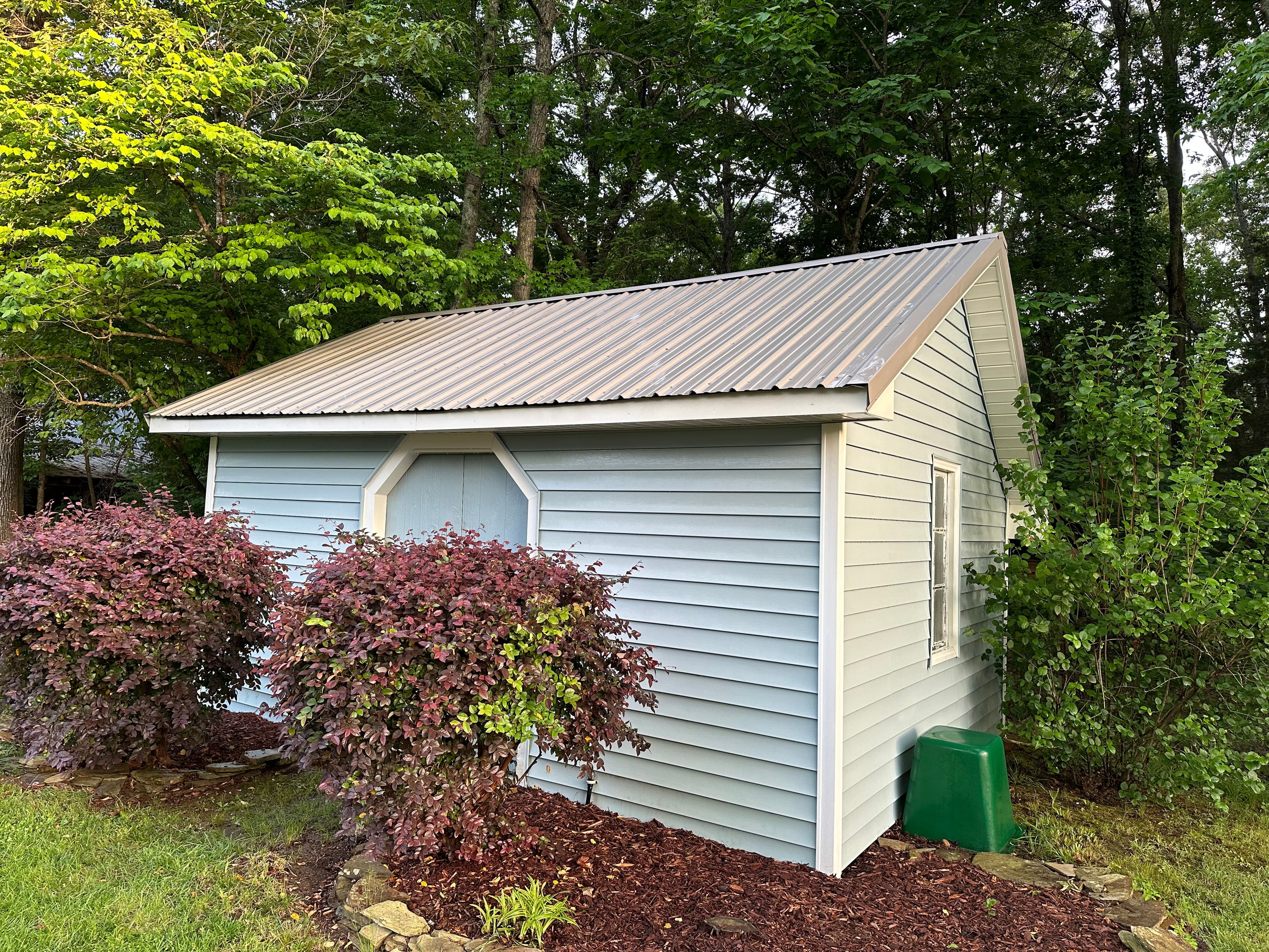 Blue shed with a metal roof surrounded by green trees and colorful shrubs in a garden setting.