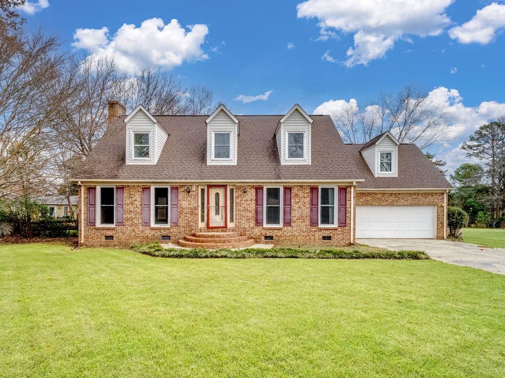 Two-story brick house with a front porch, large yard, and attached garage under a blue sky.