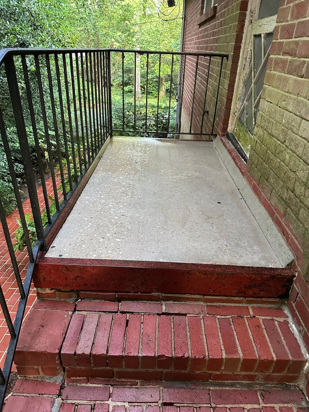 Concrete porch with red brick steps and black metal railing, surrounded by greenery.
