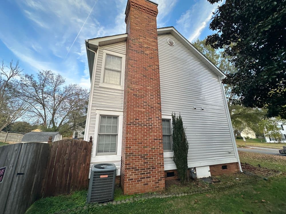 Side view of a two-story house with a brick chimney, wooden fence, and air conditioner.