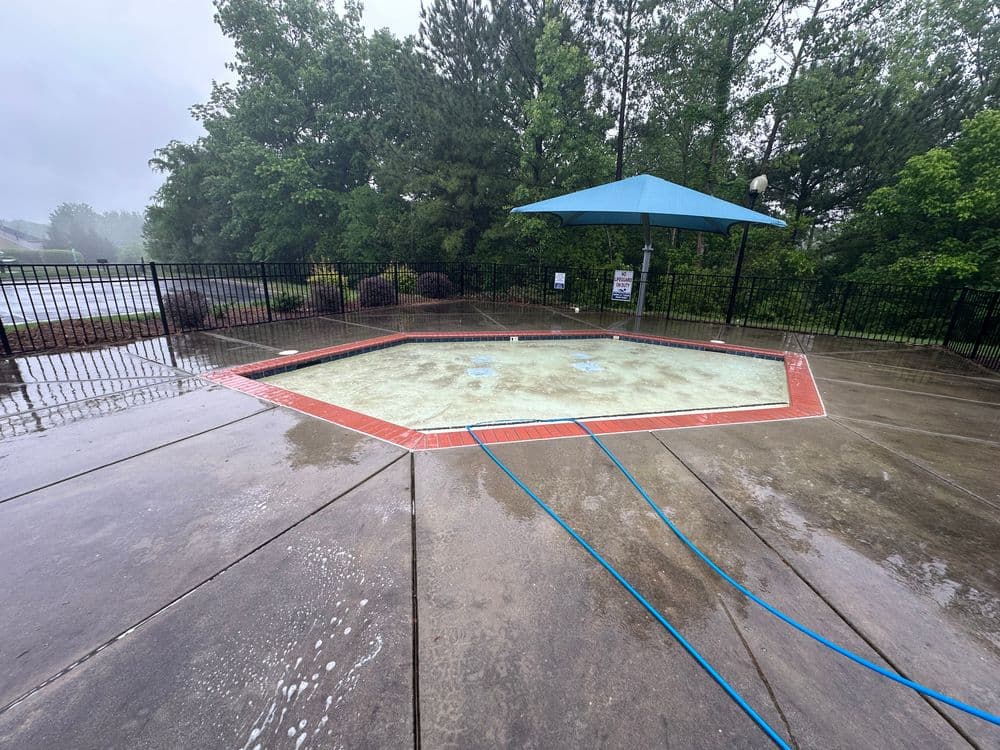 Empty pool with wet concrete deck under gray sky, surrounded by greenery and a blue umbrella.